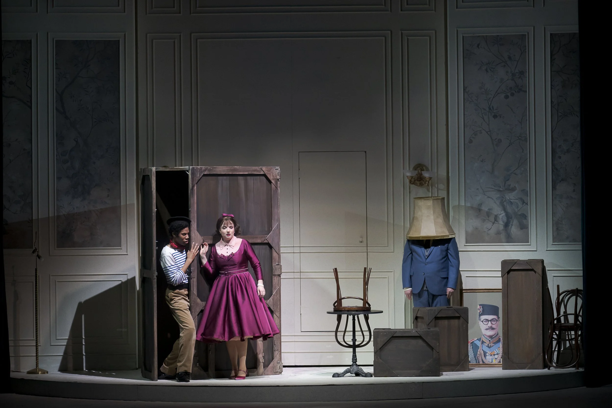 The stage from a theatrical performance featuring four actors: a woman in a pink vintage dress with a headband, a man in beige pants and a striped shirt, an actor with a costume head hidden inside a wooden box, and a person with a blue suit and a hea