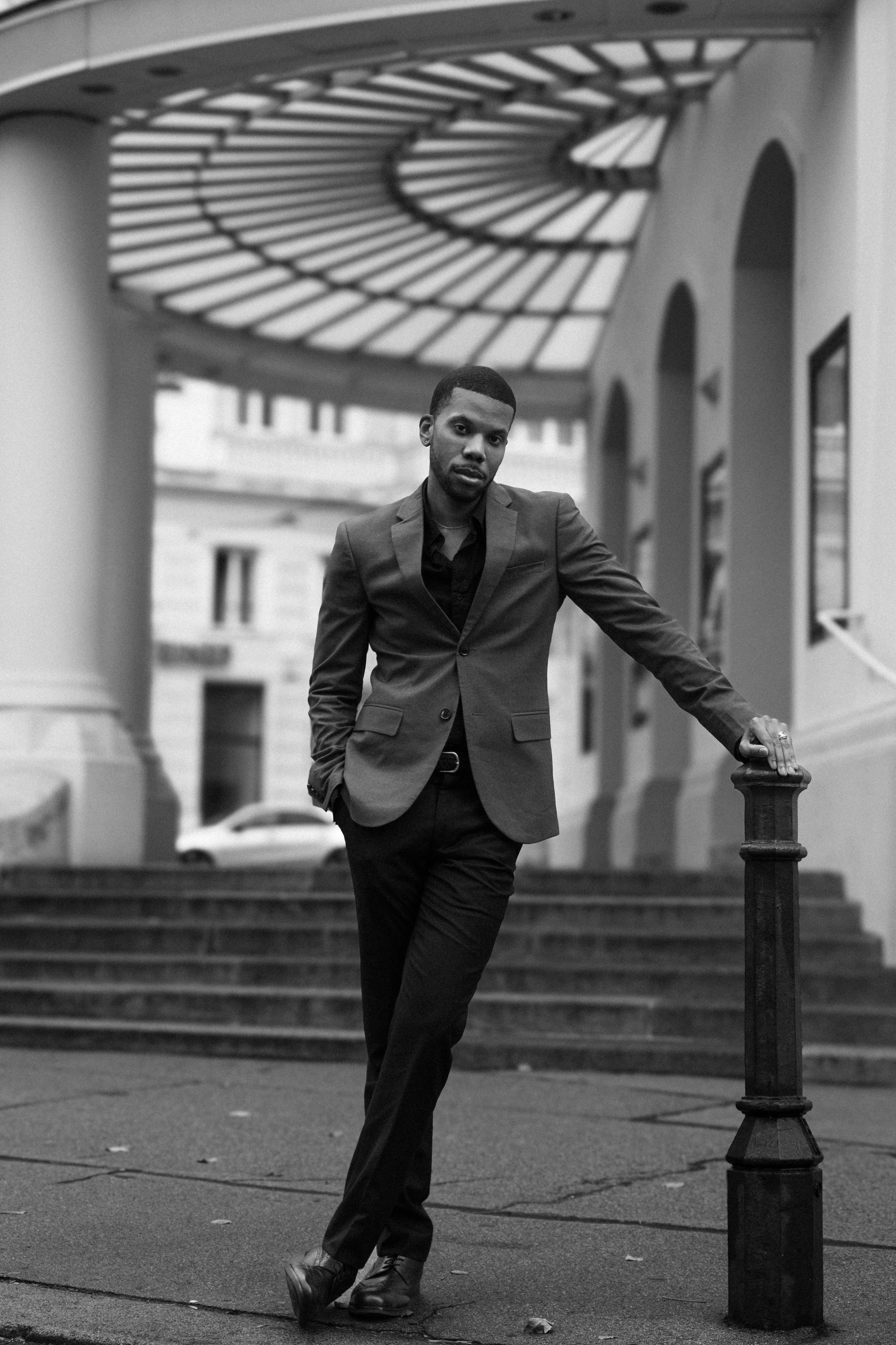 Aaron-Casey wearing a suit and dress shoes leans against a metal post on a city street. Behind him is a set of stairs and a building with arched windows. The photo is black and white.