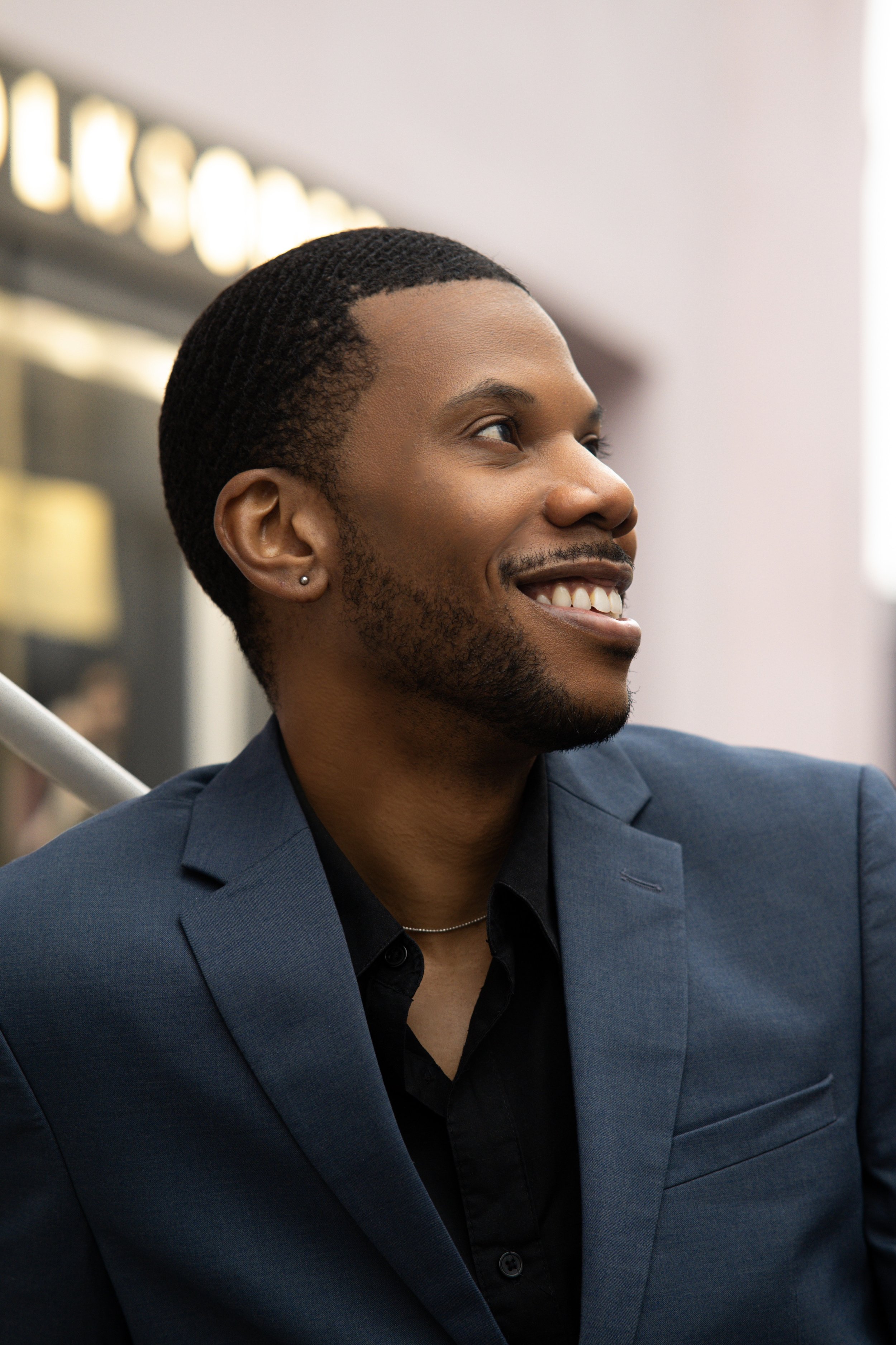 Close-up of a smiling African American man in a blue suit and black shirt, with a background blurred of lights, possibly in an indoor setting.