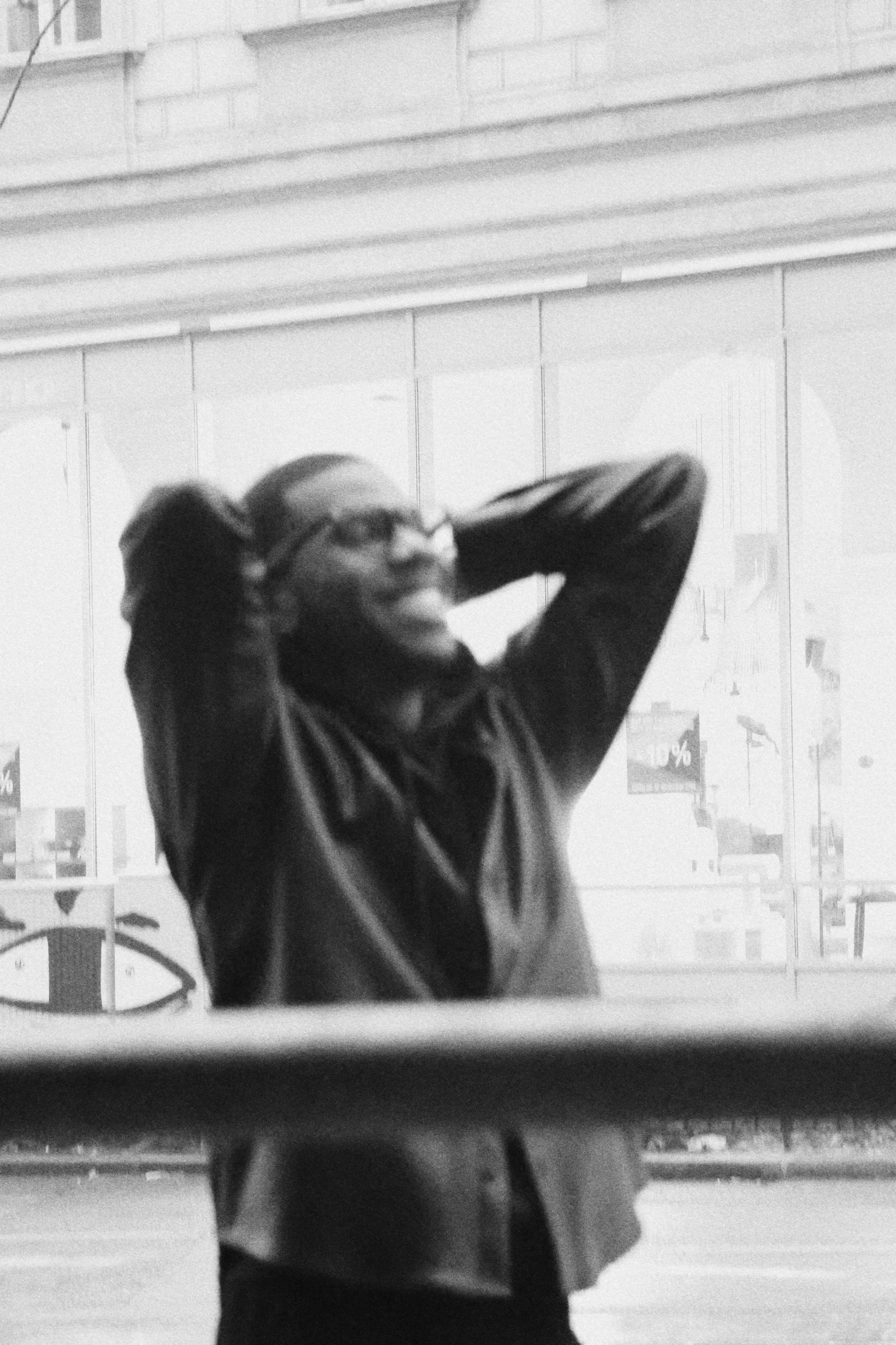 Aaron-Casey with hands behind their head, standing in front of a window display with blurry signs and objects outside.