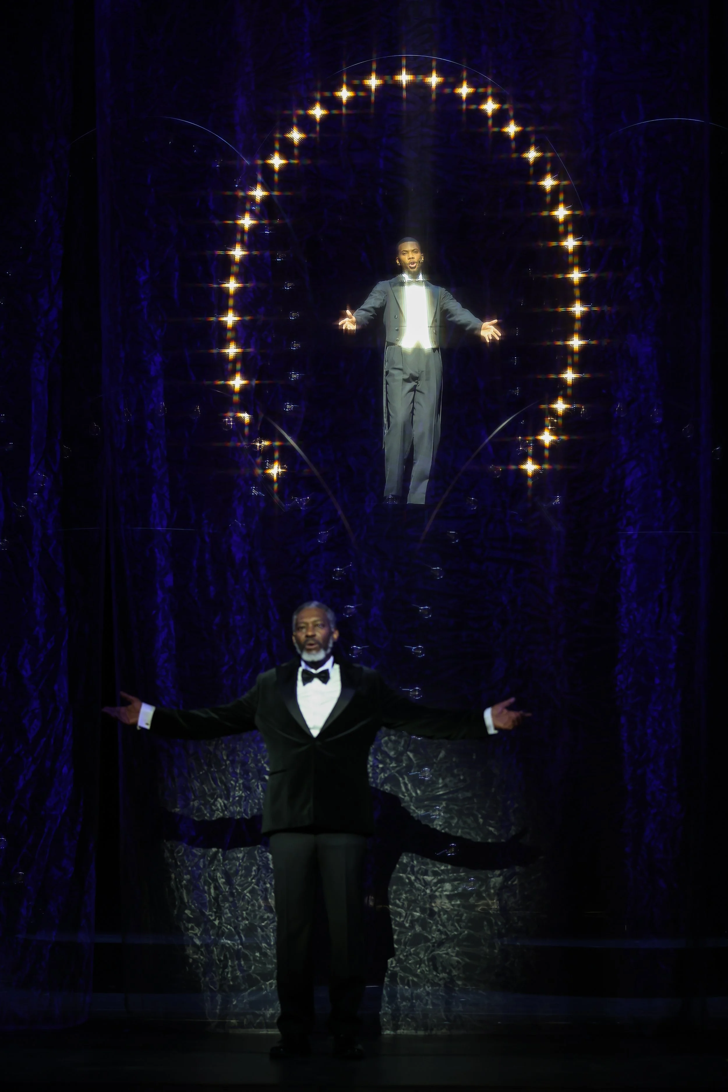 The image depicts two performers on a stage with a dark background and illuminated elements. The performer in the foreground is dressed in a tuxedo with a bowtie, standing with arms open. The performer in the background appears to be singing or speak