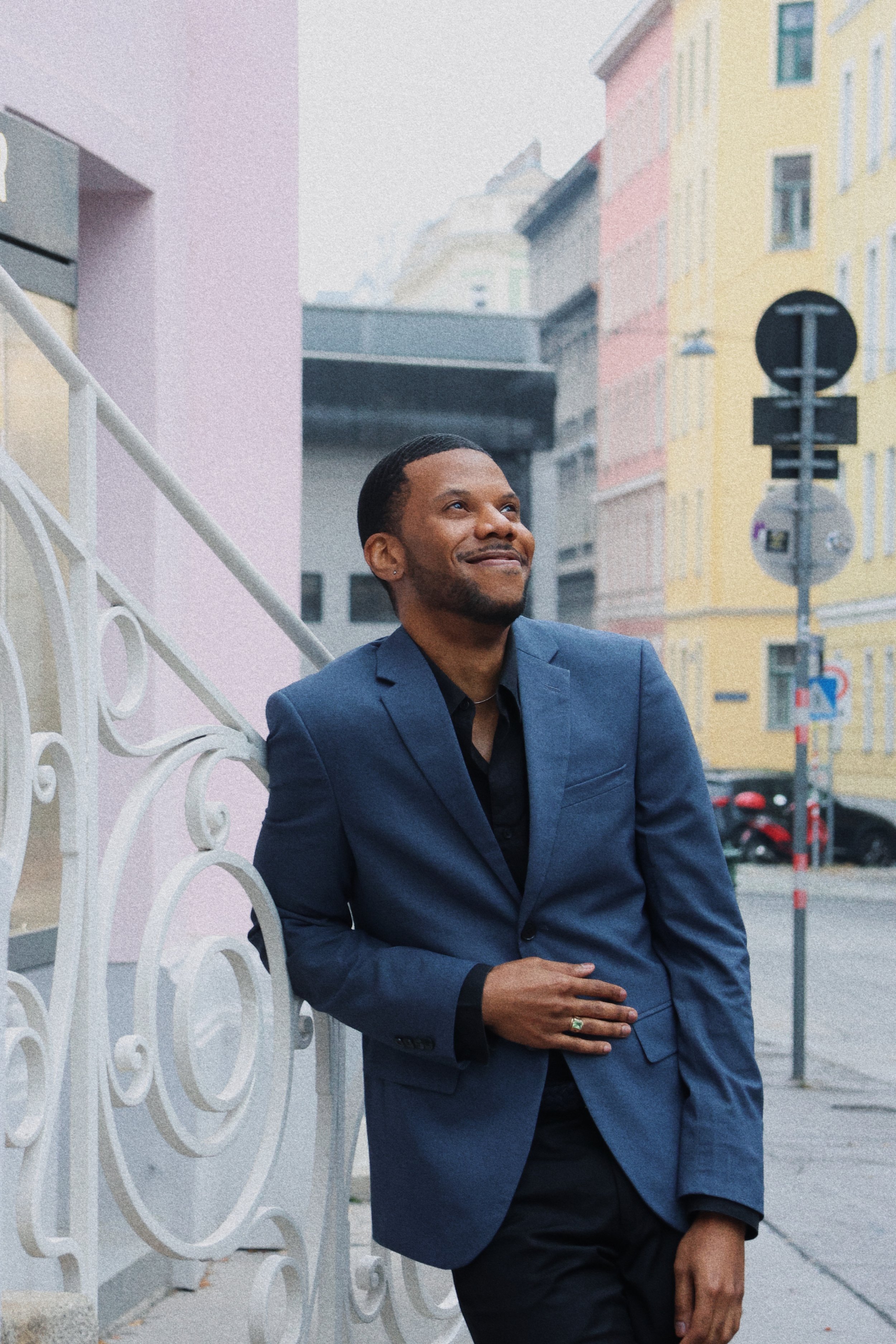 A man in a blue suit leaning against a white ornate fence on a city street, smiling and looking up with buildings in pastel colors in the background.