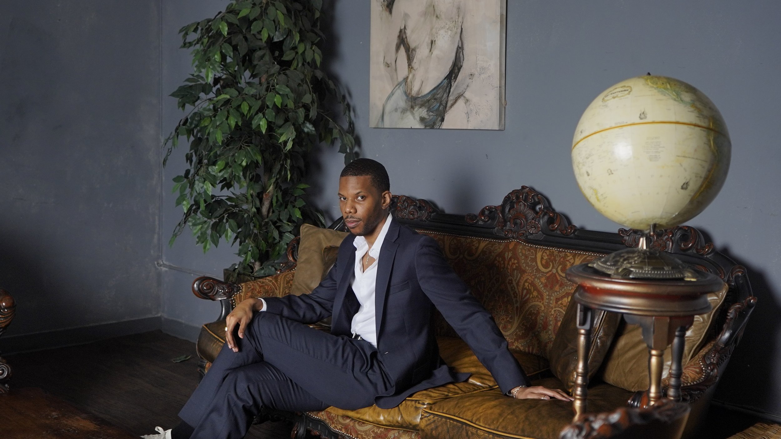 A well-dressed man with a serious expression sitting on an ornate vintage sofa in a room with dark walls, a large painting of a horse, a tall green plant, and a globe lamp on a side table.