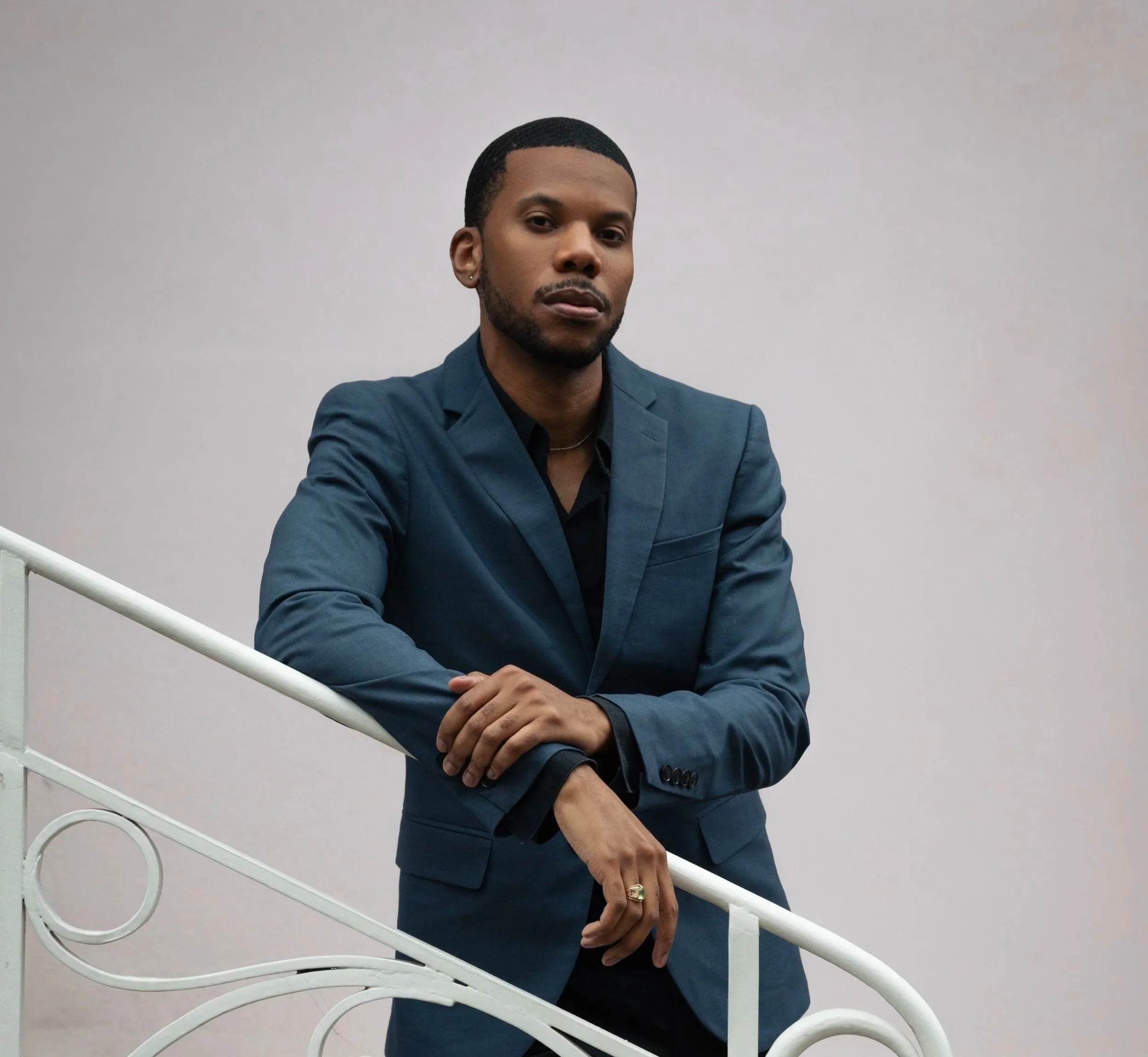 Aaron-Casey in a dark blue suit with a black shirt, standing against a plain white wall, resting his arm on a white decorative railing.