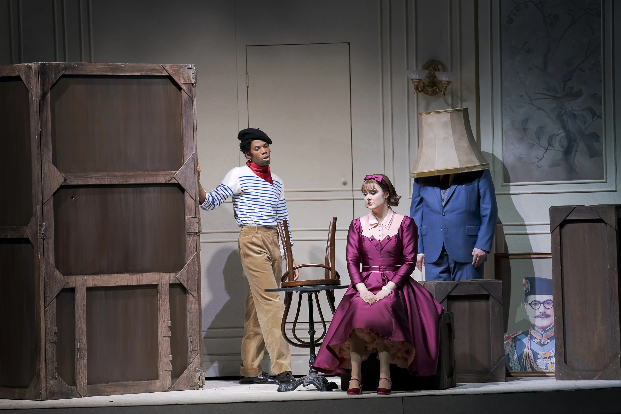 Three actors performing on stage during a theatrical play. One man is holding a large wooden box, wearing a black beret, striped shirt, and beige pants. A woman is seated in a pink satin dress with a matching bow in her hair, looking down. Another pe
