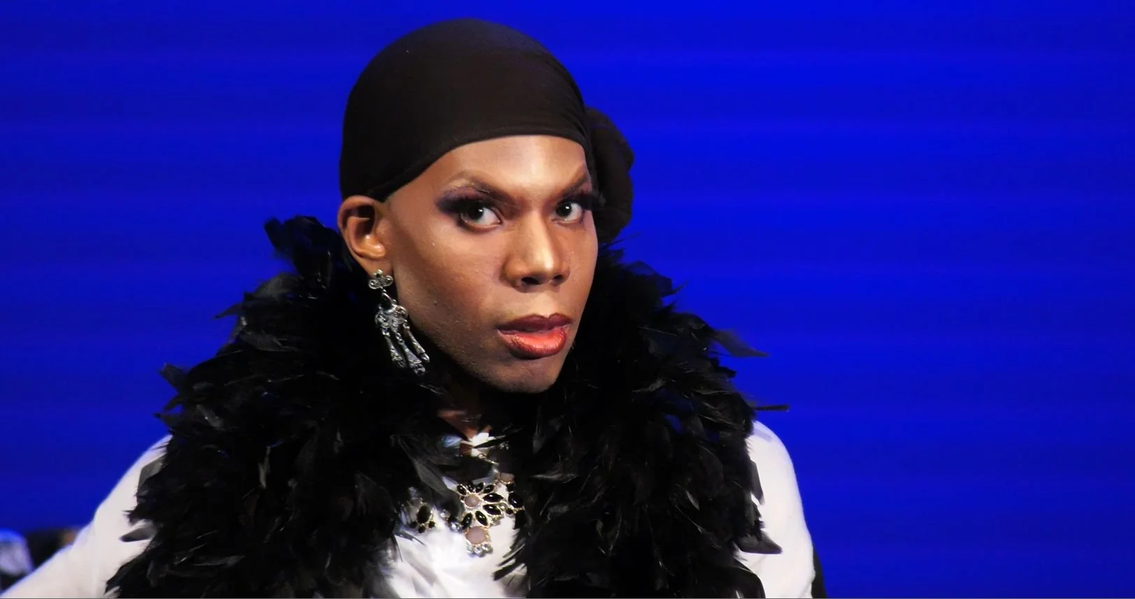 Person with makeup and earrings, wearing a black feathered accessory and black head covering, against a blue background.