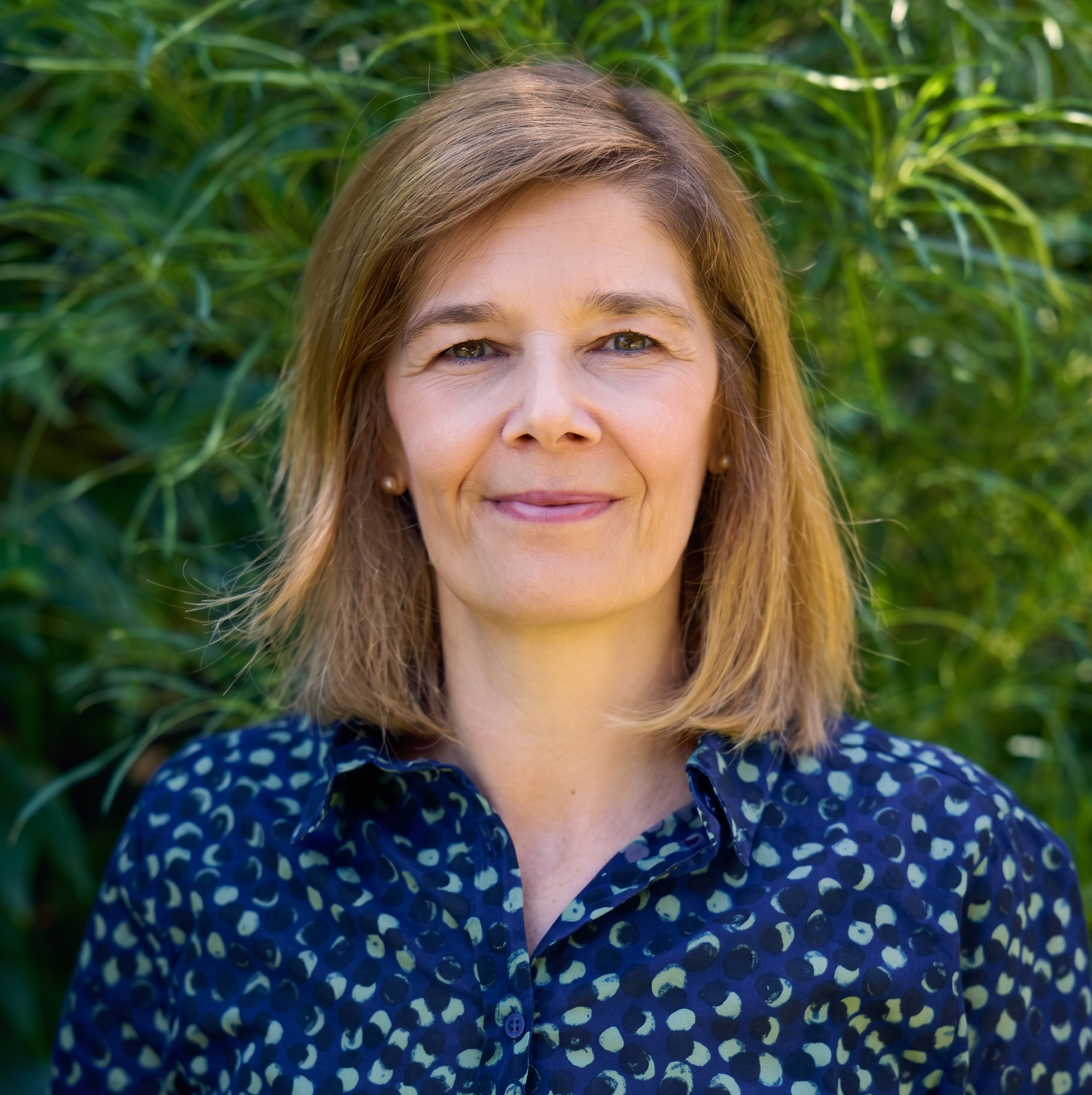 Close-up of a middle-aged woman with blonde hair, smiling, wearing a blue and white patterned shirt, outdoors with green foliage in the background.