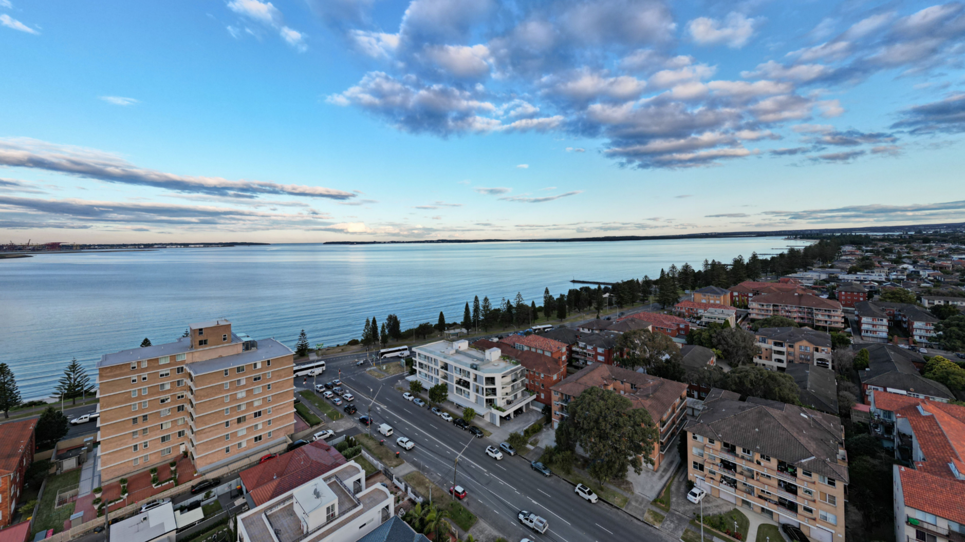 Aerial view of a coastal residential neighborhood with multi-story apartment buildings, houses, and streets lined with cars, overlooking a wide river or bay with a partly cloudy sky.