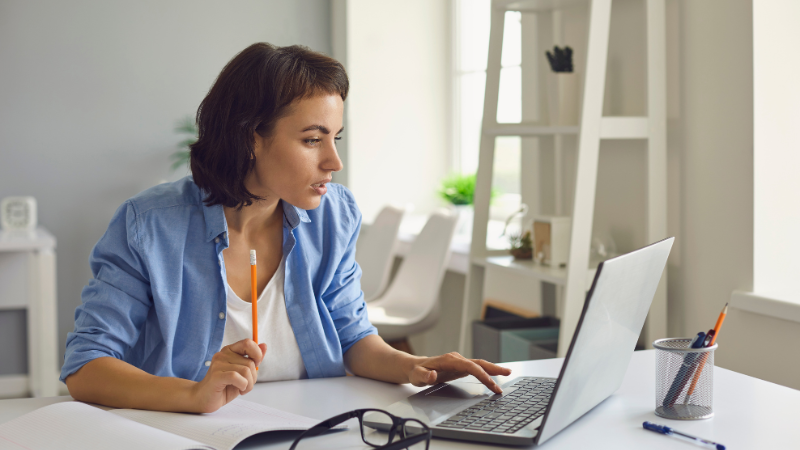 A young woman with short dark hair, wearing a blue shirt, sitting at a white desk using a laptop, holding a pencil, in a bright room with sunlight, white furniture, and a ladder shelf in the background.