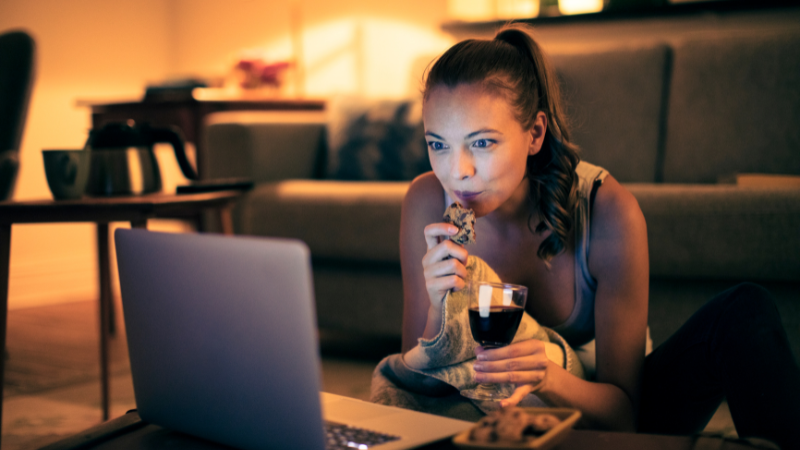 Woman watching laptop, eating cookie, holding wine glass. Cozy, dimly lit living room with sofa and warm tones, focused and relaxed mood.