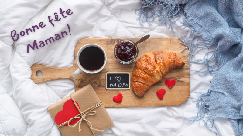 Breakfast tray with coffee, jam, croissant, and a "I ♥ MOM" note on a bed. Gift with heart, blue blanket, "Bonne fête Maman!" text.