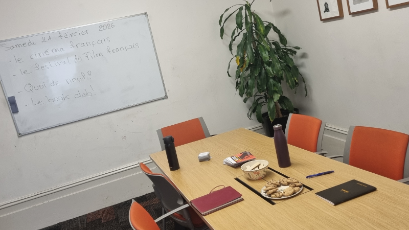 Small meeting room at Kathleen Syme Library set up for a French conversation session, with a whiteboard listing topics including le cinéma français and le book club, and a table with cookies and drinks