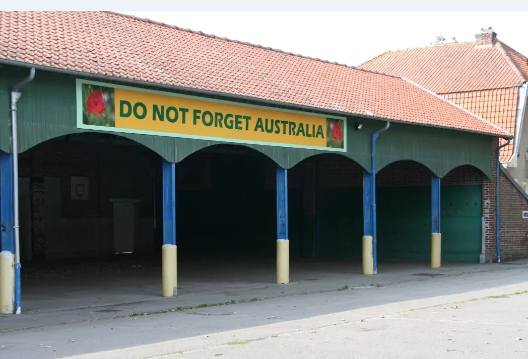Green building with a red tiled roof features a yellow sign reading "DO NOT FORGET AUSTRALIA" with poppy images. Quiet street scene.