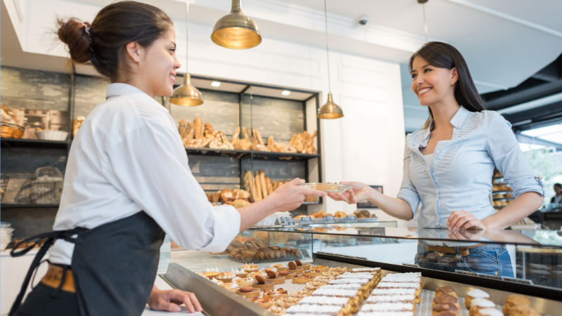 Woman in a bakery hands pastry to smiling customer. Breads displayed under warm lighting. Cozy, inviting atmosphere.