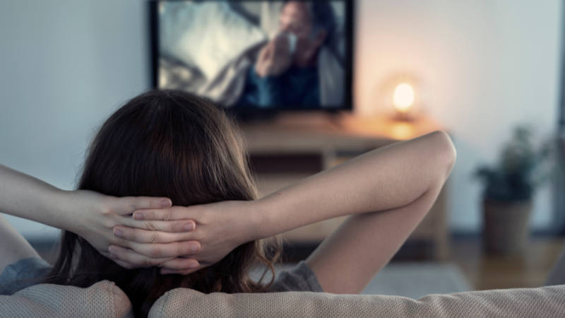A person relaxes on a sofa, watching a movie on TV. The setting is cozy with a warm lamp glow. The screen shows a person in motion.