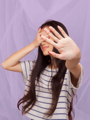 Woman in striped shirt with long hair holding hand up to camera, hiding face. Lilac background creates a mysterious mood.
