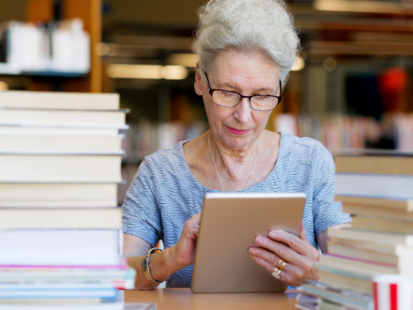 Elderly woman in glasses using tablet in library, surrounded by stacks of books. She appears focused, wearing a blue shirt.