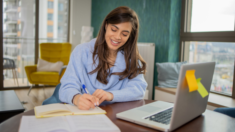 A young woman with long dark hair smiles as she writes in a notebook at her desk, with a laptop open beside her. She's in a bright, modern apartment with large windows and a city view. The mood is relaxed, motivated, and self-directed.