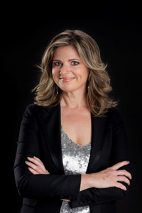 Australian television presenter and actress Julia Zemiro smiling with her arms crossed against a black background.