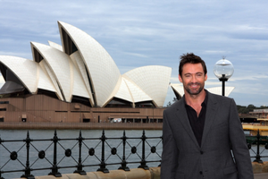 Australian actor Hugh Jackman smiling beside Sydney Harbour, with the Sydney Opera House in the background.