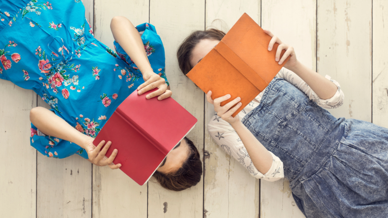 Two young women lying head-to-head on a white wooden floor, each holding a colourful book open over their faces in a playful reading pose — French book club for adults who love Francophone literature.