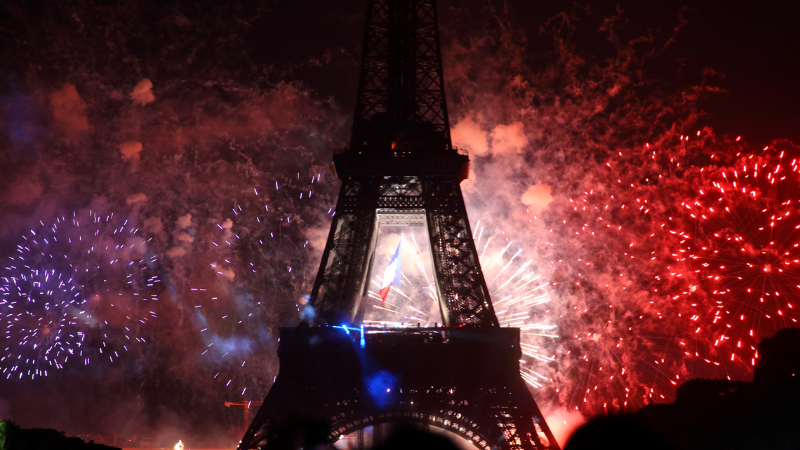 Eiffel Tower with vibrant fireworks in red, white, and blue. A French flag is visible, set against a dark night sky, creating a festive mood.