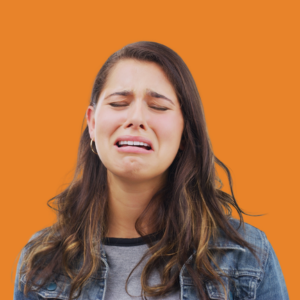 Woman with long hair in a denim jacket cries against an orange background, expressing sadness or distress.