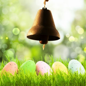 Brown bell and colorful Easter eggs on green grass with a blurred, bokeh background, conveying a festive, springtime mood.