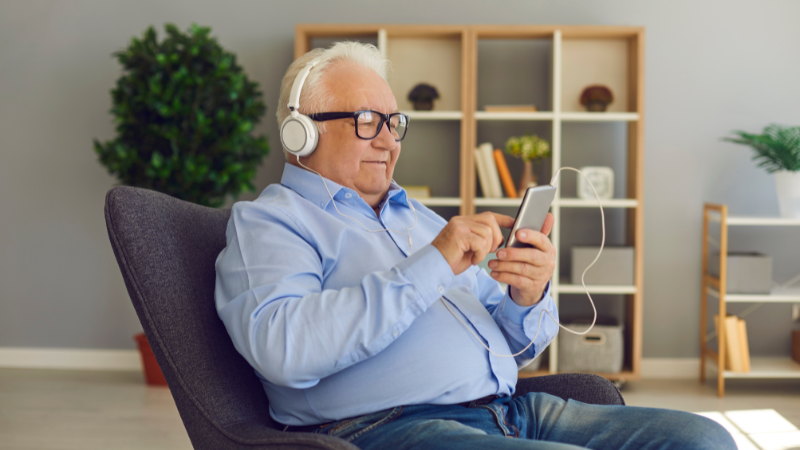 Elderly man in blue shirt sits on chair, wearing headphones, using smartphone. Shelves with plants in background; relaxed mood.