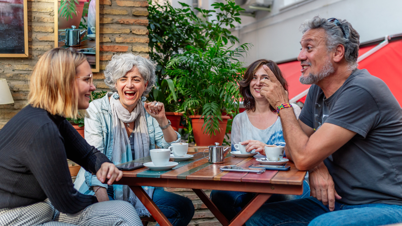 Four people laughing and talking at a cafe table outdoors, surrounded by plants. They are enjoying coffee, creating a cheerful atmosphere.