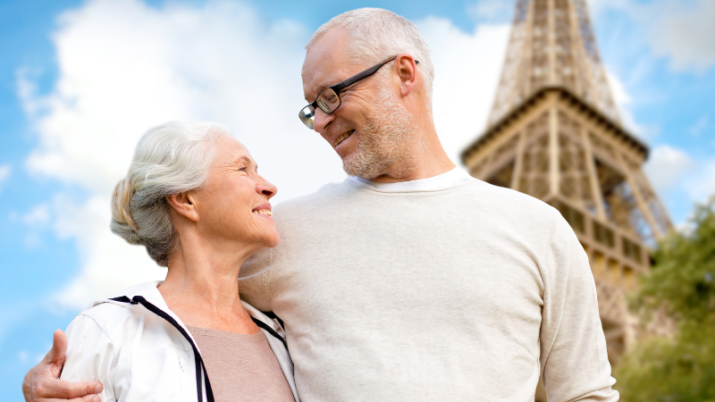 Elderly couple smiling and embracing in front of the Eiffel Tower. Blue sky with clouds. Both wear casual light-colored clothing, exuding joy.