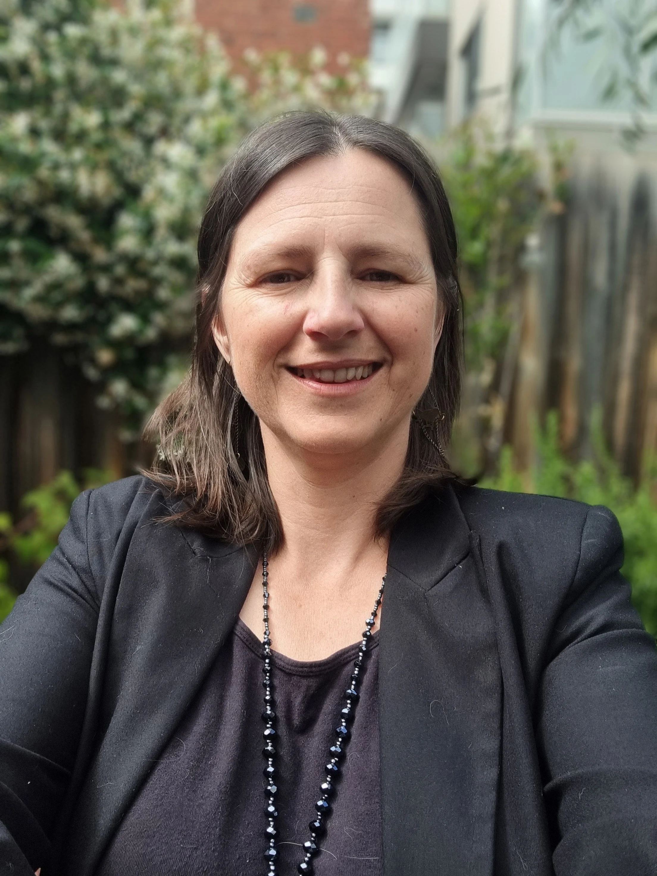 Sophie, French tutor and founder of Sophie French Tutor, smiling outdoors in a black blazer and beaded necklace — online French lessons for all ages in Melbourne