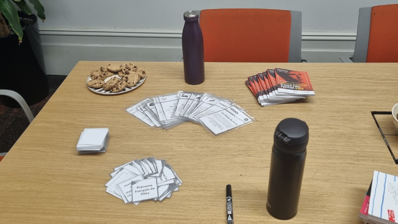 Table laid out for a French conversation group session with activity cards labelled "Prénoms français de filles", chocolate chip cookies, French Film Festival brochures, and keep cups