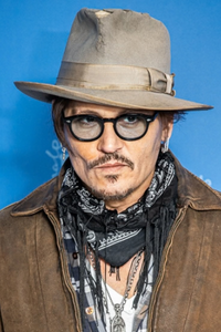 American actor Johnny Depp wearing a wide-brimmed hat, black glasses, a patterned scarf and a brown jacket at a public event.