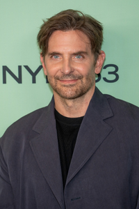 American actor Bradley Cooper smiling at a public event, wearing a dark jacket over a black shirt.
