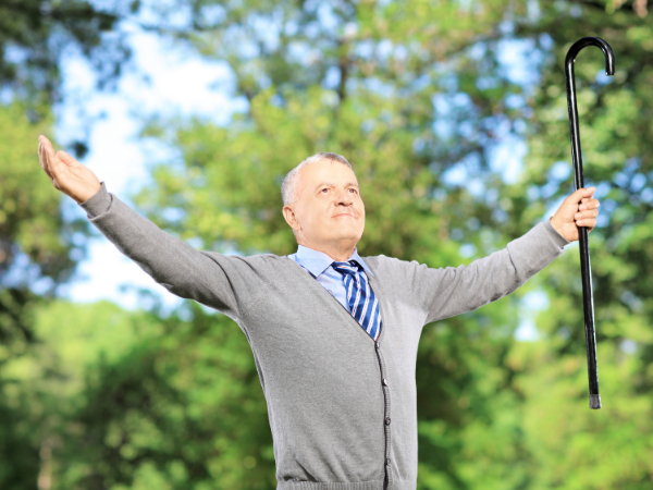 Elderly man in gray sweater joyfully raises arms with a cane in lush green park, under clear blue sky, wearing a striped tie.