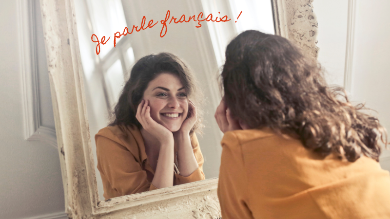 Woman smiling at her reflection in a mirror, wearing a mustard top. Text above reads "Je parle français!" in red script. Bright, cozy room.