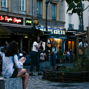 Street singer performs in a lively Paris square outside Le Requin Chagrin pub, with neon signs and seated crowd.
