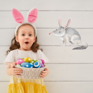 A child with pink bunny ears holds a basket of colorful eggs, surprised expression. A bilby illustration is on the white wooden wall background.