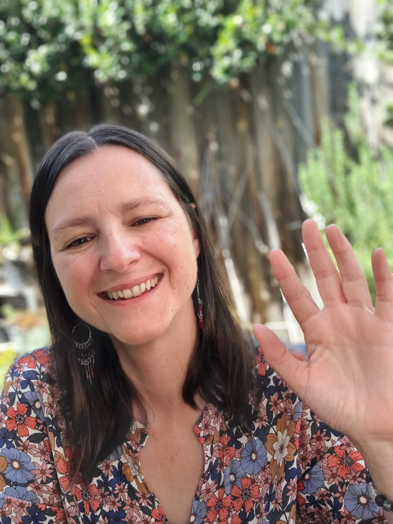 A smiling woman with long dark hair waves at the camera. She's wearing a floral dress and long silver earrings, photographed outdoors in front of a large fig tree with lush green foliage. The mood is warm and relaxed.