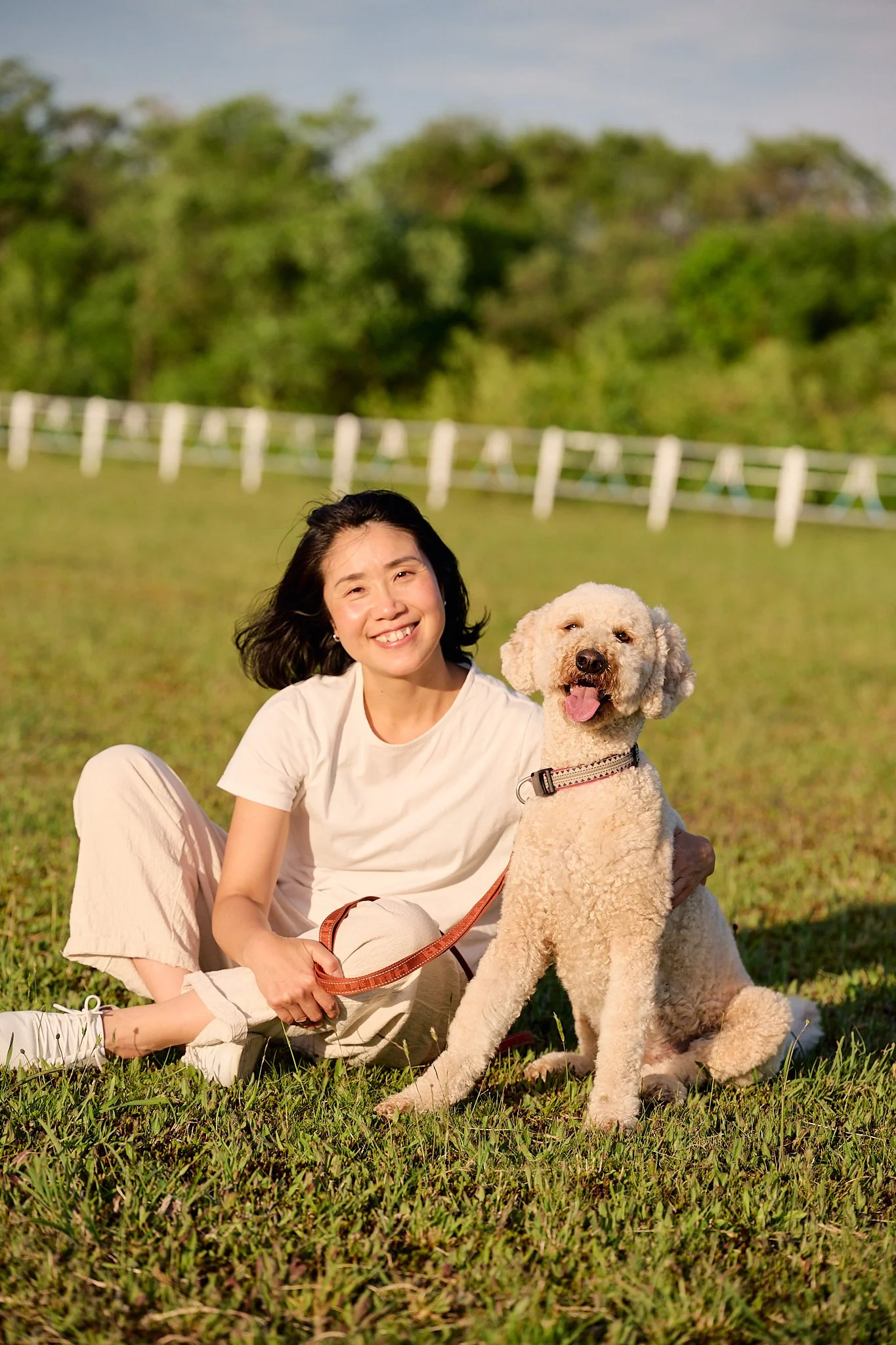 緑の草原に座る笑顔の女性とペットの犬。背景に木々と白い柵がある屋外の風景。
