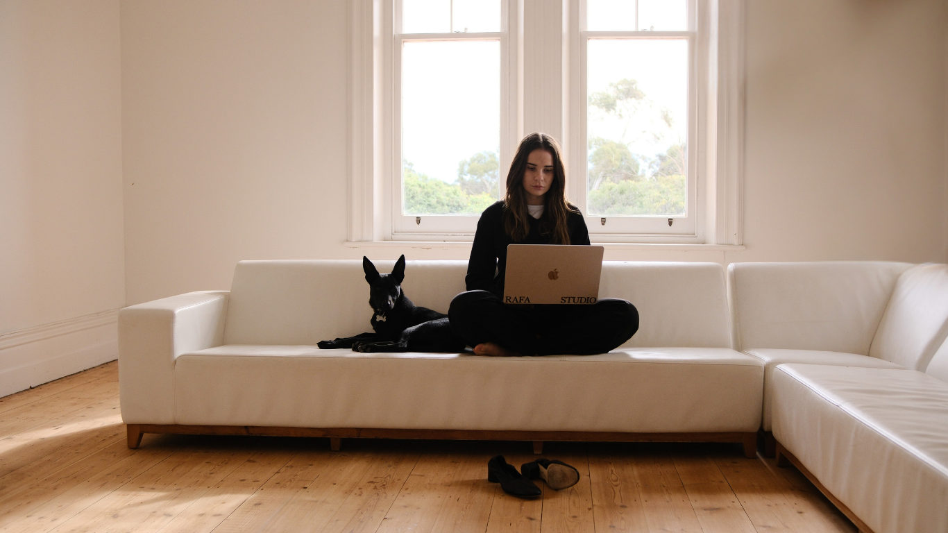 A young woman with long dark hair sitting cross-legged on a white sofa, using a laptop, with a black dog lying beside her, in a room with wooden floors and large windows revealing a view of trees outside.