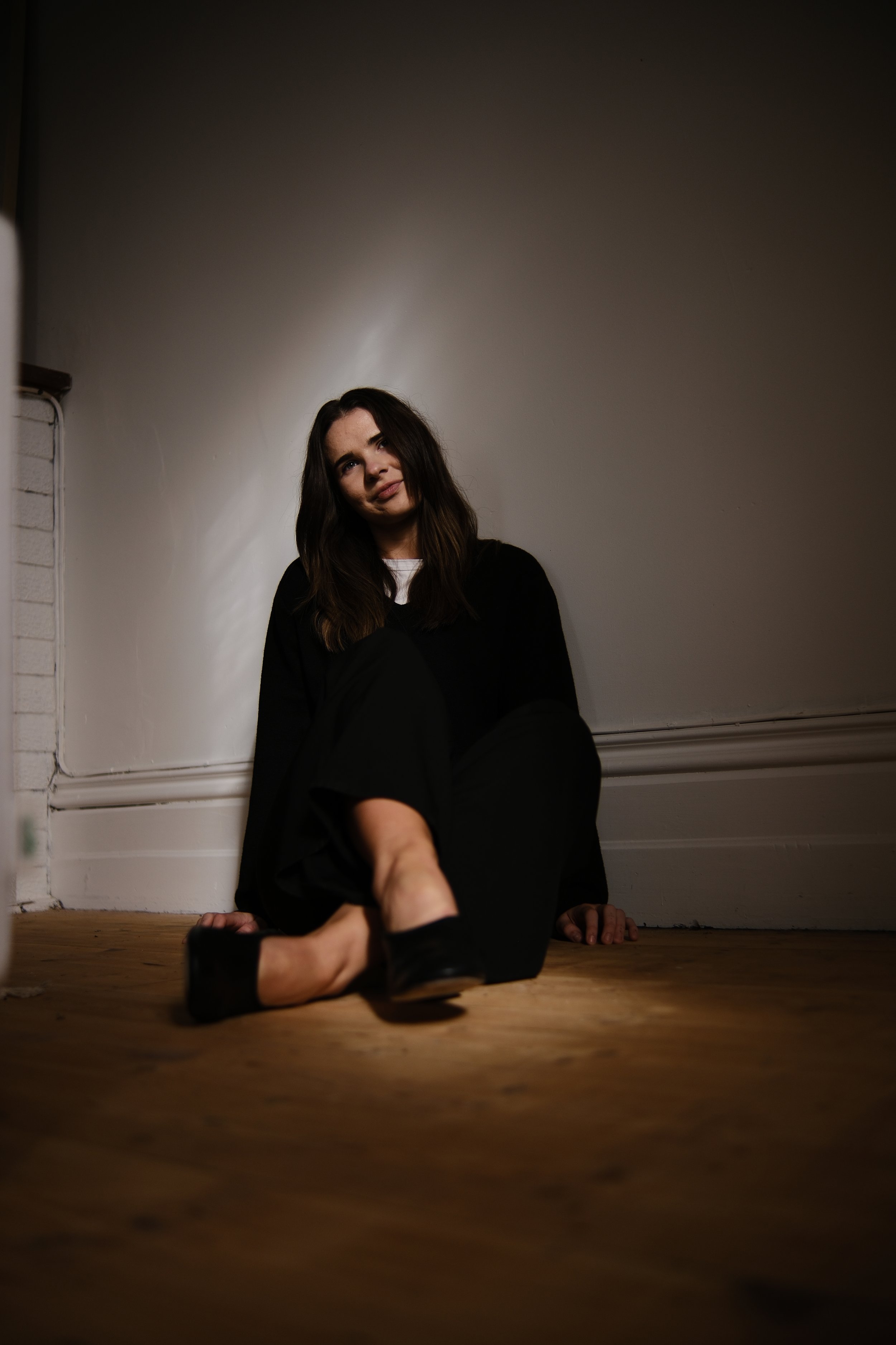 A woman with dark hair wearing a black outfit sitting on a wooden floor in a dimly lit room, with her arm extended towards the camera and a faint smile on her face.