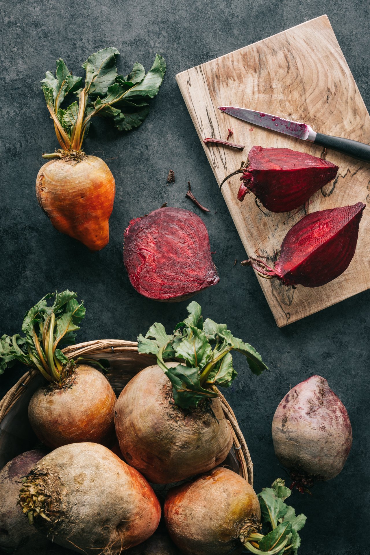 Fresh whole beets with green tops, sliced beets, and a knife on a wooden cutting board on a dark surface.