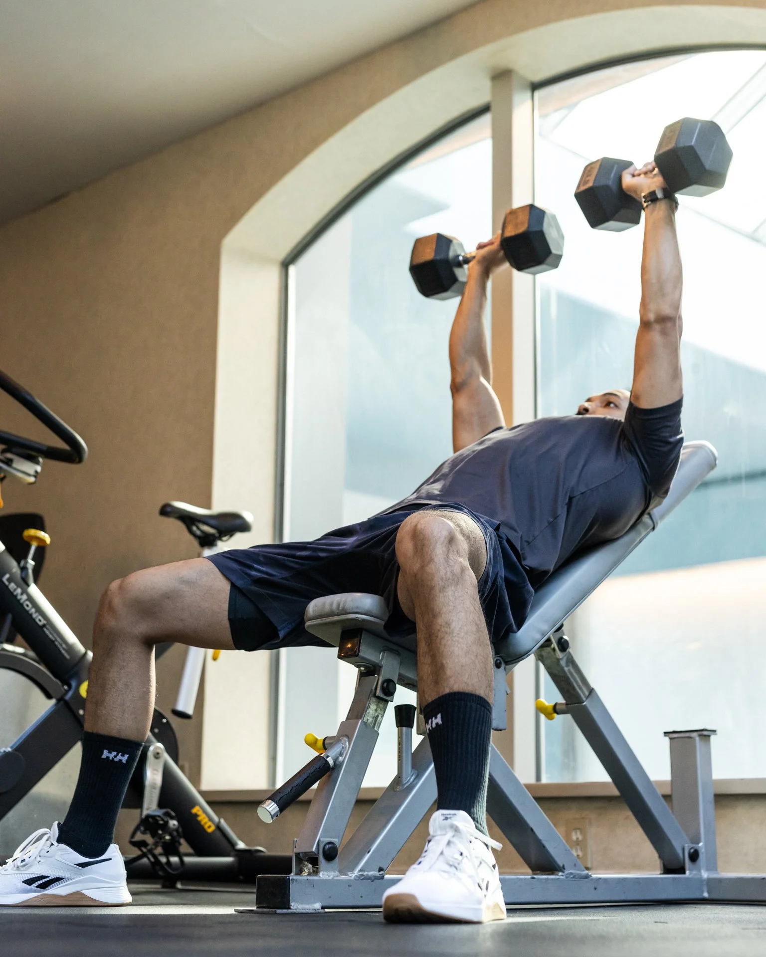 Man lying on an exercise bench lifting dumbbells in a gym with large windows.