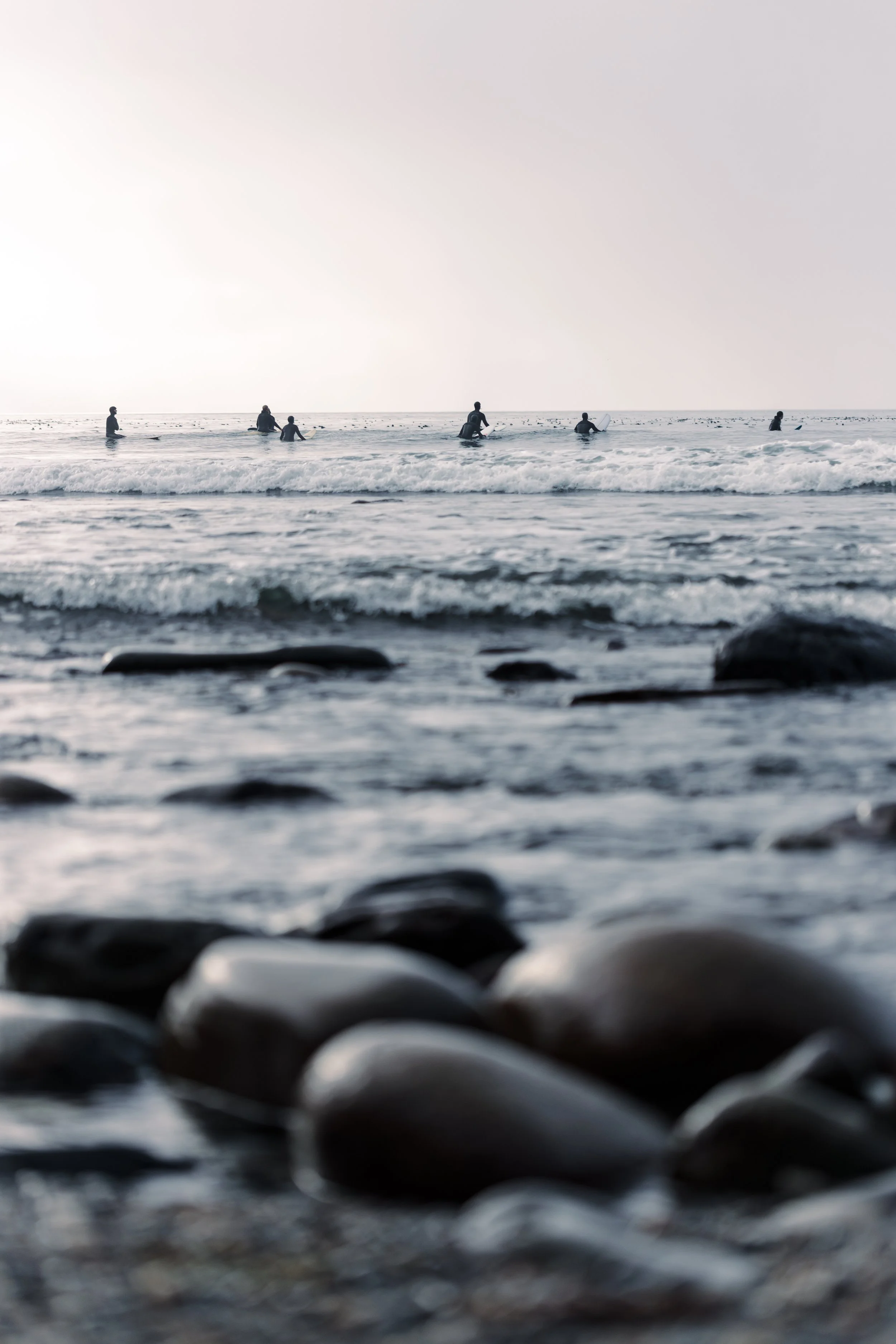 People with surfboards in the ocean at sunset or sunrise, viewed from a rocky shoreline.