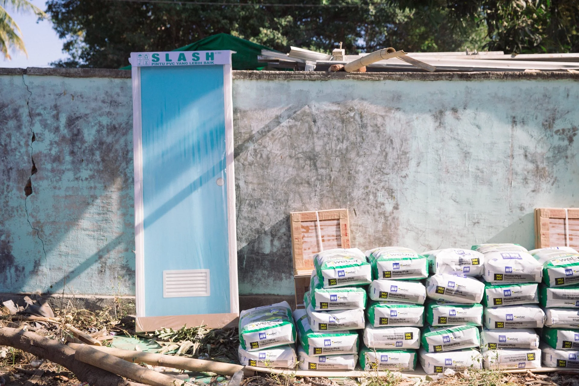 Stacks of bagged plaster placed in front of an old, weathered concrete wall with peeling paint. A small blue ventilation window and a partially visible wooden chair are also seen against the wall.