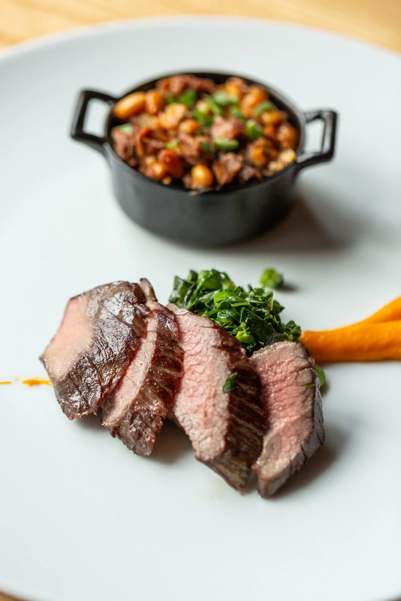 Slices of cooked steak topped with chopped herbs, served with a side of greens and a carrot on a white plate, with a small bowl of a bean and vegetable stew in the background.