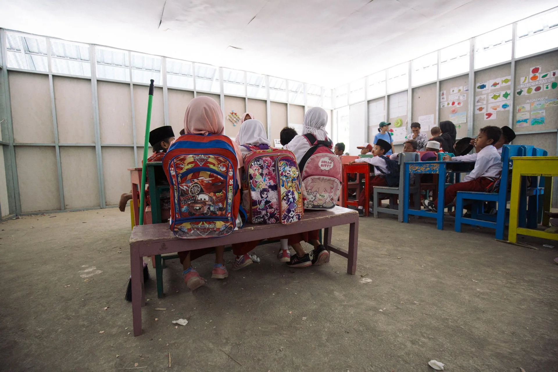 Children sitting in a classroom with backpacks on chairs and desks, and colorful drawings on the wall.