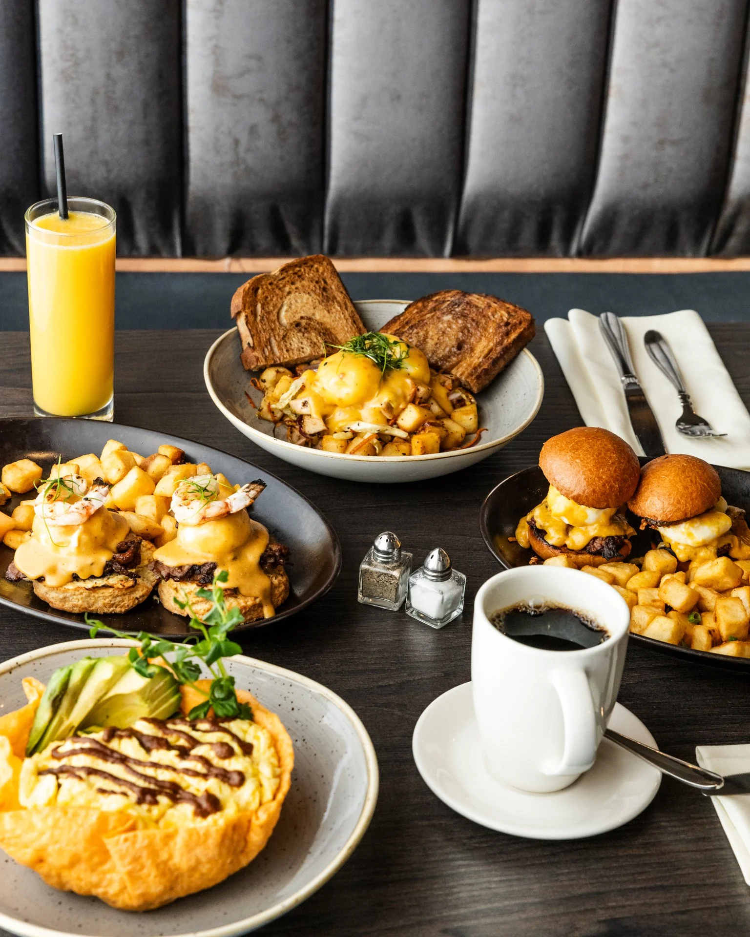 Assorted breakfast foods including eggs, toast, hash browns, muffins, coffee, orange juice, and a dish with eggs Benedict with hollandaise sauce.